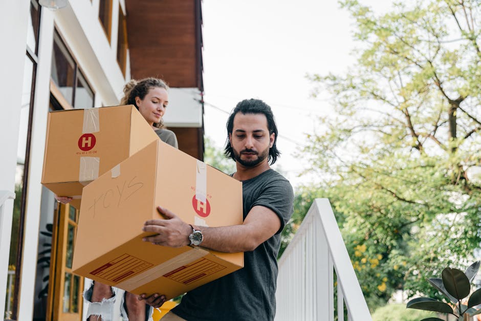 A man with shoulder-length dark hair, wearing a grey t-shirt, is carrying two cardboard boxes outdoors during a house relocation. A woman with long, curly hair, wearing a dark top, is behind him, also holding a cardboard box. The boxes are sealed with packing tape, with one showing handwritten text and red company logos. The scene takes place on a porch or entrance area with a white railing and steps, adjacent to a modern building with large windows and wooden paneling. In the background, there are green trees and a partly cloudy sky, indicating a daytime move. The man is focused on carrying himself, and the woman appears to be assisting with the move, reflecting packing and furniture transport activities common during removals. This image illustrates the packing and loading process involved in house removals, as managed by companies like Removals Harringay, in preparation for a home relocation.