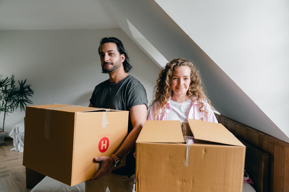 A man with shoulder-length dark hair, wearing a grey t-shirt, is carrying two cardboard boxes outdoors during a house relocation. A woman with long, curly hair, wearing a dark top, is behind him, also holding a cardboard box. The boxes are sealed with packing tape, with one showing handwritten text and red company logos. The scene takes place on a porch or entrance area with a white railing and steps, adjacent to a modern building with large windows and wooden paneling. In the background, there are green trees and a partly cloudy sky, indicating a daytime move. The man is focused on carrying himself, and the woman appears to be assisting with the move, reflecting packing and furniture transport activities common during removals. This image illustrates the packing and loading process involved in house removals, as managed by companies like Removals Harringay, in preparation for a home relocation.