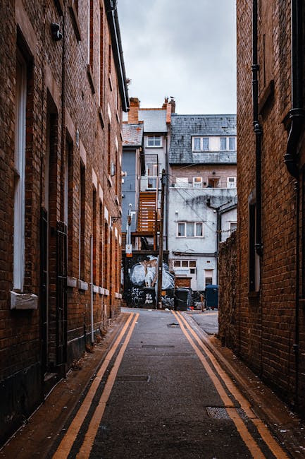 A narrow urban alleyway between two brick buildings in a residential area, featuring a tarmac surface with yellow double lines along the edges. At the far end, there is a small set of wooden stairs attached to the back of a grey building, with a few windows and a door visible. Two black waste bins are positioned at the base of the stairs. The alley is illuminated by overcast natural light, and the scene suggests a typical backstreet environment suitable for residential removals and home relocation activities carried out by Removals Harringay, as the space is cleared and ready for furniture transport or moving logistics.