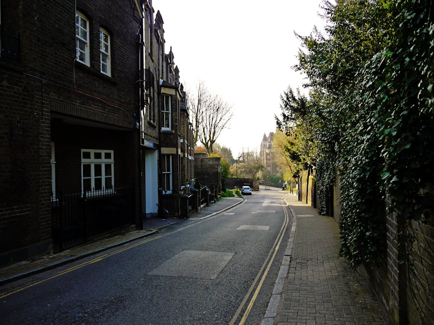 A narrow residential street with a slight incline seen during daylight, featuring dark brick terraced houses on the left side with white-framed windows and small front gardens enclosed by black metal fences. The street is lined with a high, dense hedge and trees on the right side, casting shadow over the paved sidewalk made of concrete slabs. The road surface is asphalt with dual yellow lines along the curb, and a few parked cars are visible further down the street. The scene offers a typical urban setting suitable for house removals and furniture transport, with ample space for loading and unloading, consistent with a guide for relocating homes in Harringay. Removals Harringay occasionally manage such logistics within these streets for packing, moving, and transporting furniture, supported by equipment like trolleys and moving blankets, although none are visible in this specific image.