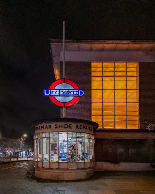Nighttime exterior view of Harringay Green Lanes station showing the illuminated London Underground roundel sign mounted on a pole, with the station's name partially visible below it. To the left of the sign, a small, rounded brick building with a curved glass window displays a shoe repair shop named 'MARMAR SHOE REPAIRS', with various tools and footwear visible inside. The background features a brick building with large, vertically aligned windows emitting warm yellow light, and part of the street scene is faintly illuminated by nearby streetlights. The scene appears quiet, with no visible pedestrians or moving vehicles, capturing a calm, indoor lighting ambiance suitable for a house relocation and moving services context connected to Harringay.
