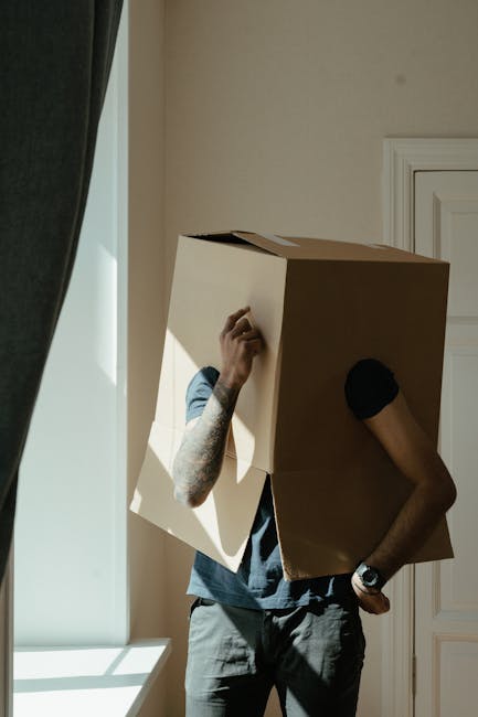 A person wearing a short-sleeved grey T-shirt, with visible tattooed arms, is carrying a large cardboard box during a home relocation process inside a room. The individual is positioned near a window with natural daylight illuminating the scene and a dark curtain partially visible on the left side. The person's right arm is bent, gripping the box from underneath, while their left hand rests on their hip, which is visible through the person's posture. The room features a white wall with a white door or panel to the right. The box appears to be new and sturdy, suitable for packing household items. This scene is representative of the packing and carrying stage involved in house removals and furniture transport, as handled by professional services such as Removals Harringay, as featured on their website for guidance on hidden charges and moving logistics.