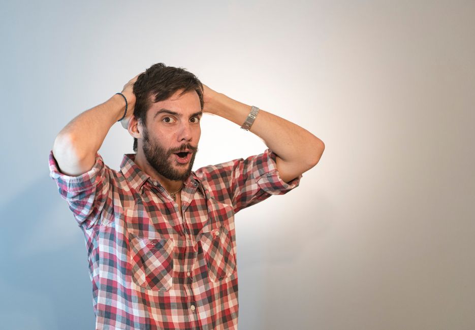 A man with dark brown hair and a beard, dressed in a red, white, and blue checkered shirt, is standing indoors against a plain, neutral background. He has a surprised or confused expression on his face, with his mouth slightly open and eyebrows raised. He is holding his head with both hands, fingers interlocked behind his head, as if expressing frustration or disbelief. Partially visible on his wrist is a silver watch. The setting appears to be a home or an office, with no visible furniture or other objects in the frame. This image illustrates the concept of house removals or relocation stress, aligning with the context of hidden removals charges and moving services offered by Removals Harringay, as the man’s expression can symbolize the common surprises or concerns during a home relocation, especially related to costs or moving logistics.