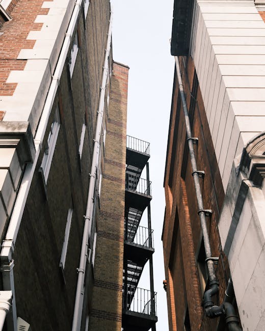 A view looking upward between two narrow buildings with brick and stone facades, showing a metal fire escape staircase attached to the side of one building, extending vertically with multiple landings