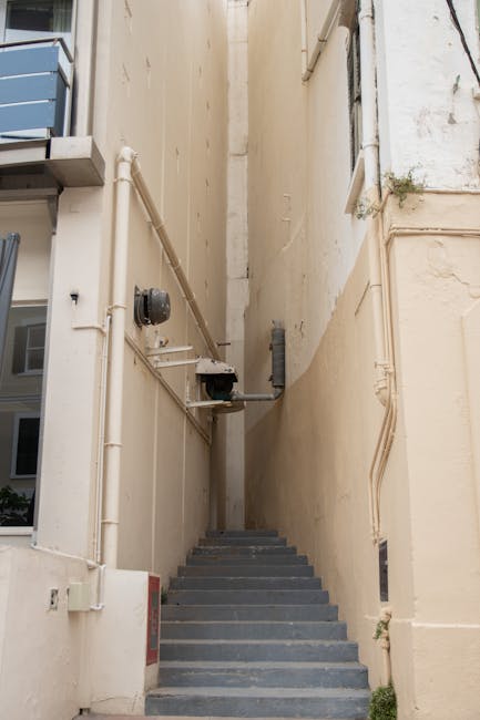 A view looking upward between two narrow buildings with brick and stone facades, showing a metal fire escape staircase attached to the side of one building, extending vertically with multiple landings