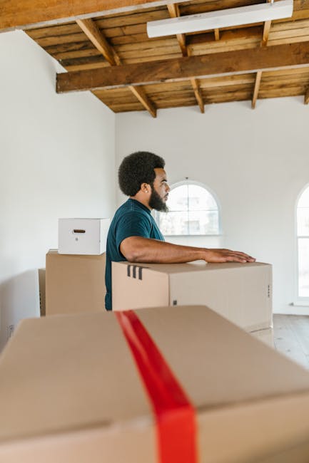 A man with dark curly hair and a beard, wearing a blue shirt, stands inside an unfurnished room with white walls and a wooden ceiling with exposed beams. He is placing his hands on top of a large cardboard moving box, which is sealed with clear packing tape. Several other similarly sized boxes, some with tape and some without, are stacked nearby on the floor and on a piece of furniture. The room features two arched windows that let in natural light, illuminating the space. The scene captures the process of home relocation, with the man preparing or organising boxes for a furniture transport or packing and moving service, consistent with professional removals provided by Removals Harringay. The environment appears clean and minimally furnished, emphasizing the focus on the boxes and the loading process within the interior space as part of the scheduled move or move planning.