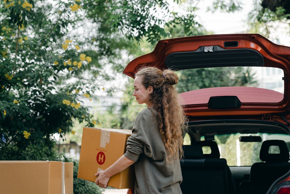 A woman with long, curly hair tied back in a bun is loading a cardboard box onto a vehicle during a home relocation or packing process, with the rear hatch of a red car open behind her. She is dressed in a grey sweater and is holding the box securely with both hands. The vehicle is parked on a street or driveway, with other boxes visible nearby, some wrapped in plastic or tape for protection. In the background, there are lush green trees with yellow flowers and a partly cloudy sky. The scene illustrates the loading process typical of furniture transport or removals services, such as those provided by Removals Harringay, supporting tips for parking and loading on moving day as seen on the webpage N4 removals tips for parking and loading on moving day, Harringay.