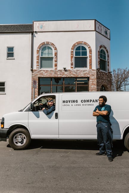 A white moving van parked in front of a modern, two-storey residential building with a brick and white façade under a clear blue sky. Two men are present: one inside the vehicle, visible through the open driver’s side window, appears to be a mover or homeowner engaging in a conversation, while the other stands outside the van with arms crossed, wearing a blue shirt and jeans, observing the loading process. The van bears the logo and text for a local moving company, indicating its role in home relocation and furniture transport. The scene captures a typical packing and loading phase of a house removal, with the vehicle positioned near the property’s entrance for efficient access. Reflecting the logistics involved in professional removals, the setting emphasizes the arrangement of packed boxes, furniture, or packaging materials that may be inside the van. This image is relevant to the services provided by Removals Harringay, focusing on house removals and moving operations in the Harringay Ladder area.