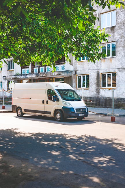 A white cargo van parked on a residential street beneath a tree with lush green leaves casting shadows on the pavement. The van is positioned next to an older apartment building with multiple windows and a concrete exterior. The vehicle appears to be part of a home relocation process, with no visible loading or unloading activity in progress. Nearby, a metal fence runs along the sidewalk, and the area is illuminated by daylight, indicating clear weather. The setting reflects an urban environment suitable for conducting furniture transport or packing and moving operations, with the van ready for part of a larger moving service provided by Removals Harringay, which specialises in house removals and household logistics.