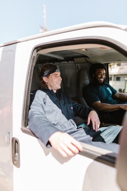A white moving van parked in front of a modern, two-storey residential building with a brick and white façade under a clear blue sky. Two men are present: one inside the vehicle, visible through the open driver’s side window, appears to be a mover or homeowner engaging in a conversation, while the other stands outside the van with arms crossed, wearing a blue shirt and jeans, observing the loading process. The van bears the logo and text for a local moving company, indicating its role in home relocation and furniture transport. The scene captures a typical packing and loading phase of a house removal, with the vehicle positioned near the property’s entrance for efficient access. Reflecting the logistics involved in professional removals, the setting emphasizes the arrangement of packed boxes, furniture, or packaging materials that may be inside the van. This image is relevant to the services provided by Removals Harringay, focusing on house removals and moving operations in the Harringay Ladder area.