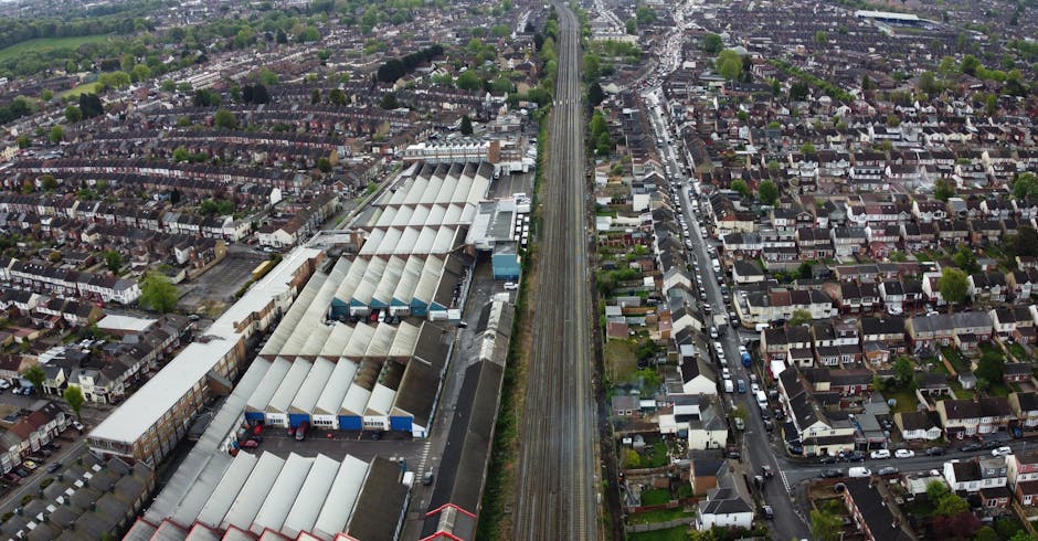 Aerial view of a residential area in Harringay showing rows of terraced houses with rooftops, alongside a railway line running vertically through the centre of the image. To the left, there is a large industrial warehouse complex with a series of interconnected buildings featuring curved, corrugated metal roofs, some with visible loading bays and parked delivery trucks. The right side depicts narrow streets lined with parked cars and tightly packed houses, with small gardens and greenery interspersed throughout. The lighting is natural, indicating daytime, and the image captures the urban environment where furniture transport, packing, and loading processes are likely underway as part of house removals or home relocation efforts. The scene emphasizes the proximity of residential and commercial properties, relevant to the services offered by Removals Harringay.